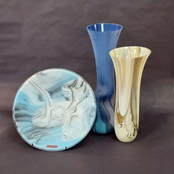 Round blue glass plate beside a tall glass blue vase and cream brown vase against dark grey background